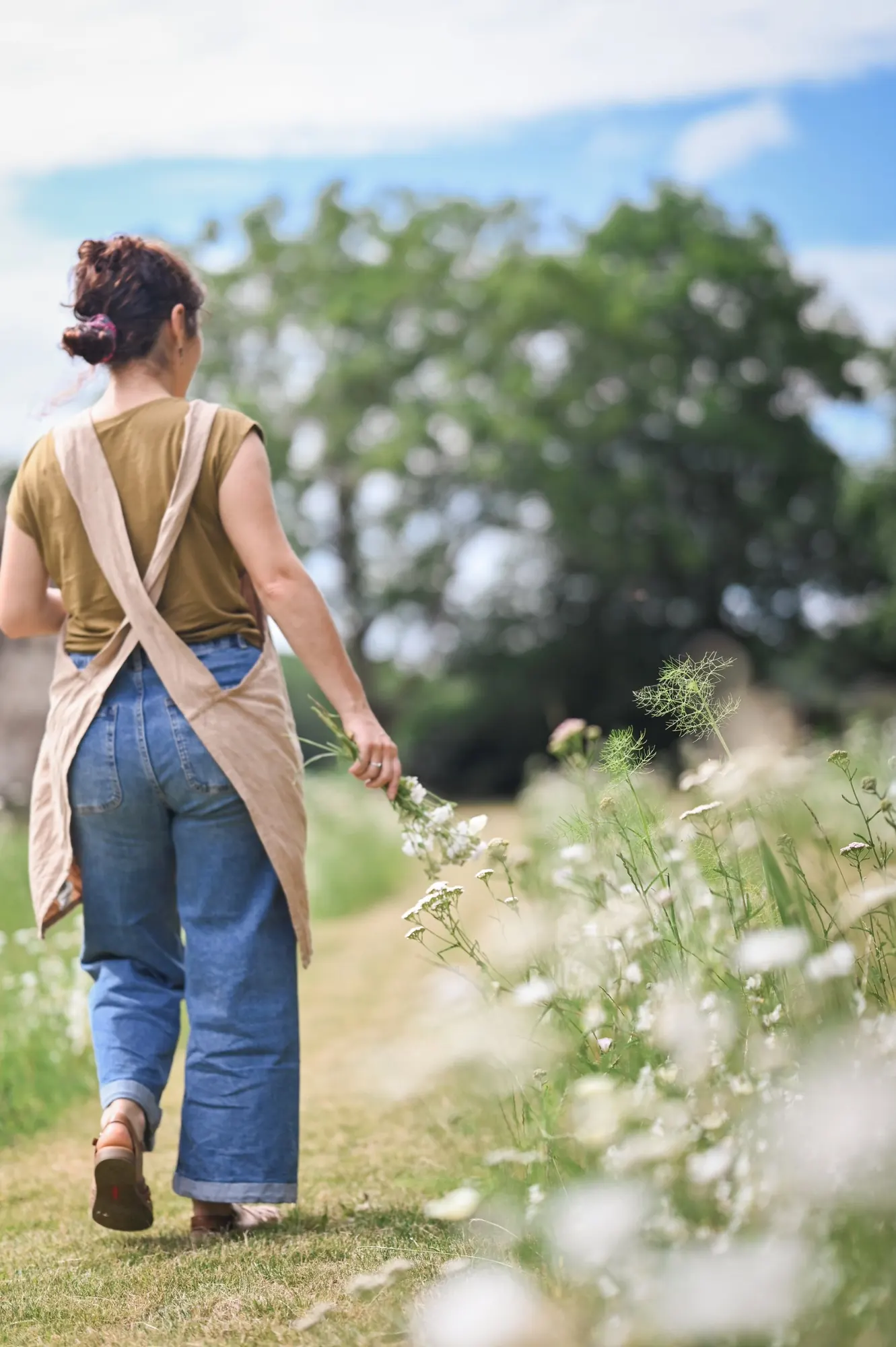A woman in a linen apron walking along a mown path with white flowers